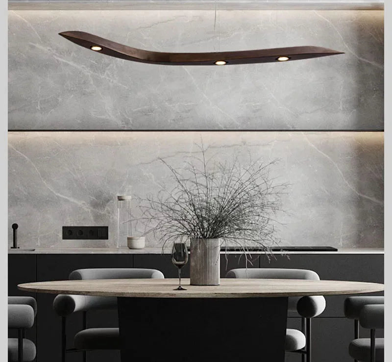 A modern dining area with a round table, gray chairs, a vase of dry branches, and the Spiral Wood Pendant Light brings warmth and elegance to the gray marble wall backdrop.