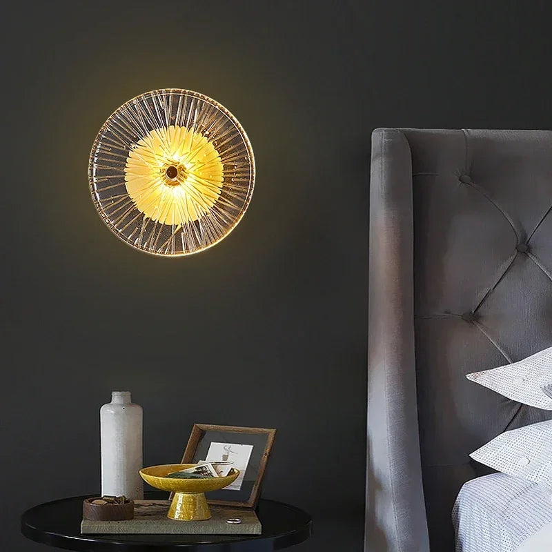 The Amber Deco Wall Sconce emits warm light above a black round side table with decor, beside a gray upholstered bed set against a dark wall.