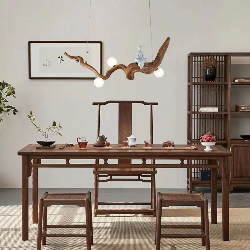 A minimalist dining room showcases a natural wood table with two chairs and a shelf. Above, the Zen-Inspired Wooden Pendant Light resembles branches, casting a soft glow on wall art. Tea sets and fruit bowls complete the zen aesthetic.