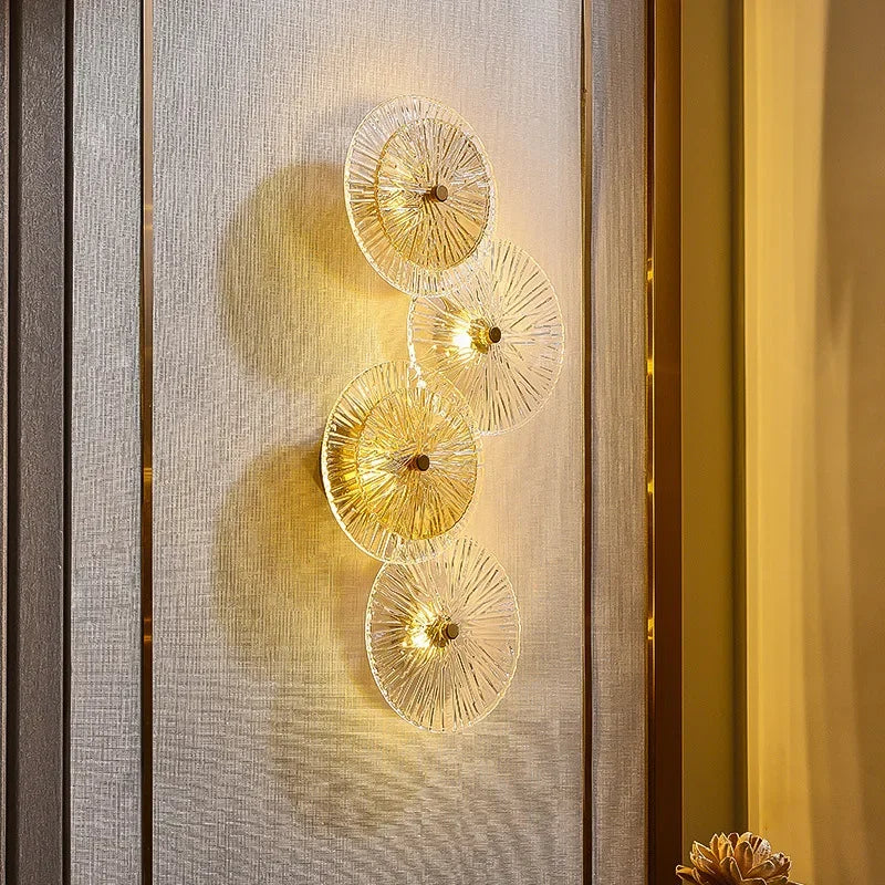 The Amber Deco Wall Sconce is a wall-mounted lamp featuring four vertically arranged glass discs, casting a warm yellow light onto a textured beige wall.