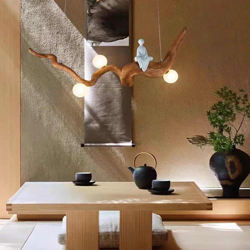 A minimalist tea room features a natural wood table, tea set, and bonsai. The Zen-Inspired Wooden Pendant Light resembling a branch with orbs enhances the Zen aesthetic, illuminating a small sculpture and the wall scroll.