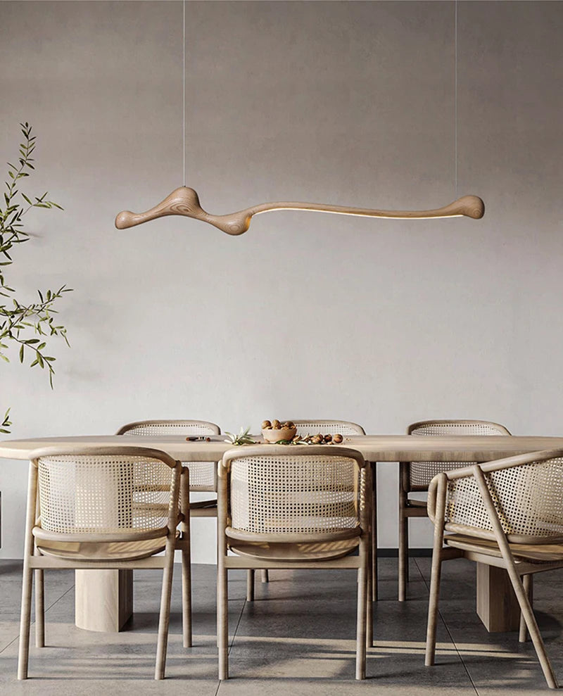 A minimalist dining room with a light wood table, six woven chairs, the Tideform Biomorphic Pendant Light overhead, and a potted plant set against a plain, neutral-colored wall.