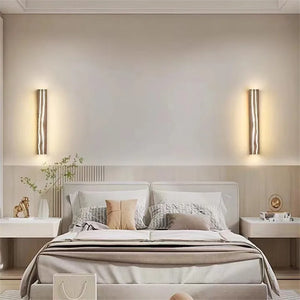 A modern bedroom features a neatly made bed with neutral bedding, flanked by minimalist Rectangle Wood Wall Lights. The decor embraces a wooden aesthetic, complemented by small plants and objects on bedside tables, creating a serene ambiance.