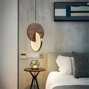 A modern bedroom features a Dual Disc Pendant Light above a small wooden side table with a plant. A framed abstract artwork adds sophistication, while gray and white bedding completes the scene.