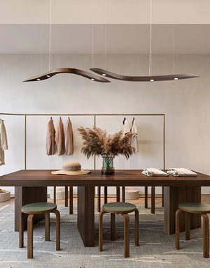 A modern room with a wooden table, three stools, a vase of pampas grass, the Spiral Wood Pendant Light overhead, clothes on a rack, and neutral decor accents.