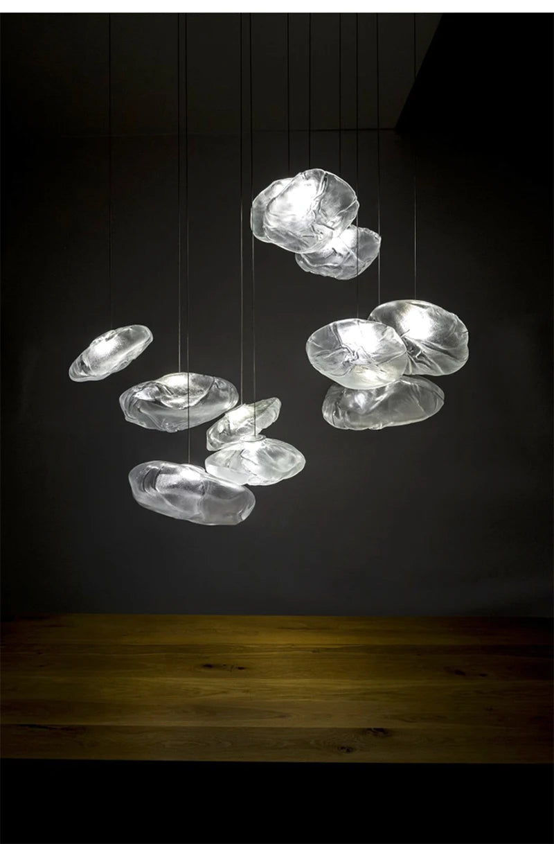 The Luxe Cloud Glass Pendant Light, resembling a floating cloud, is suspended above a wooden surface and softly glows against a dark background.