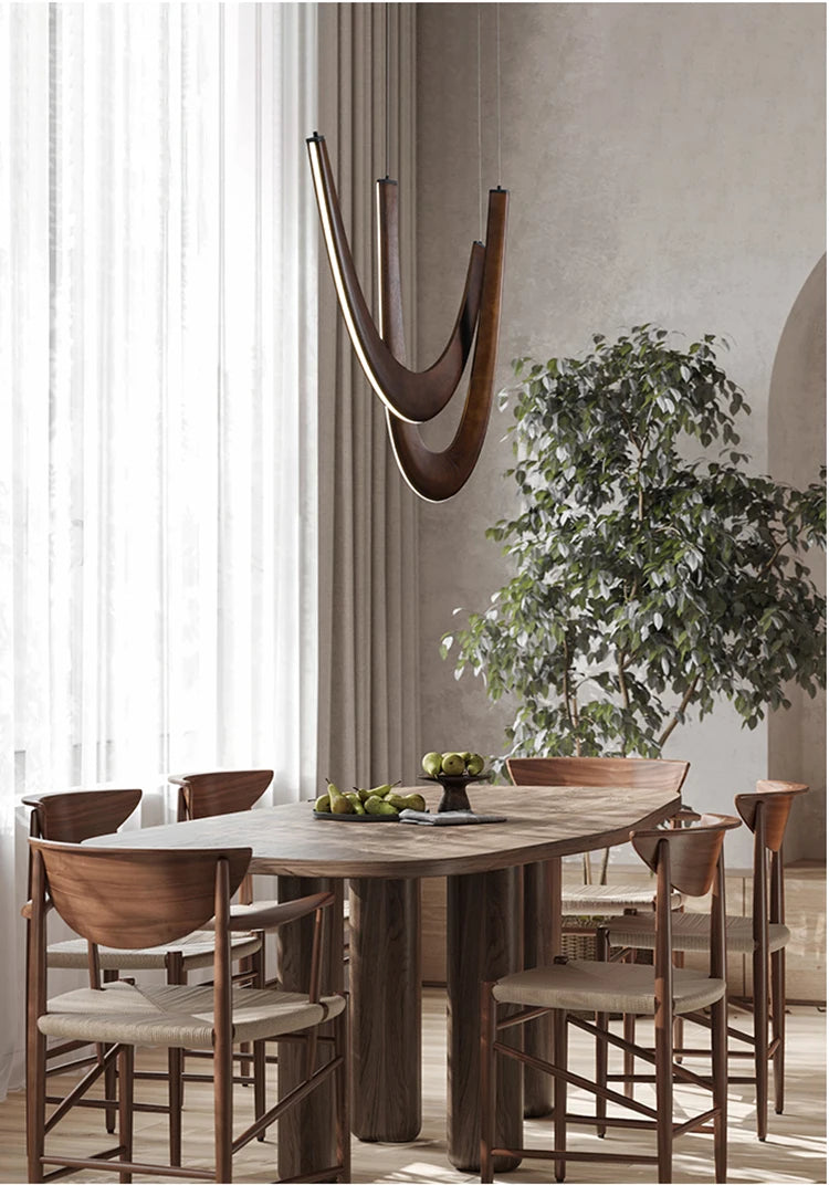 A modern dining room with a wooden table, six chairs, a Double Curve Wood Pendant Light, a potted tree, and sheer curtains that let in natural light.