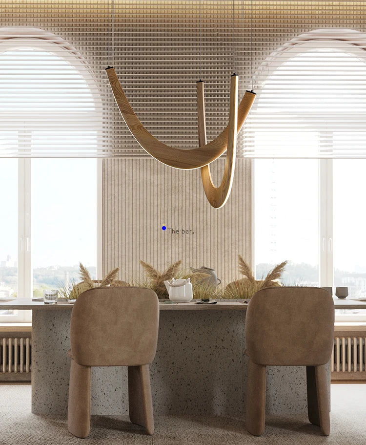 Modern dining area featuring two beige chairs, a stone table, decorative dried grass, and the Double Curve Wood Pendant Light hanging in front of large arched windows with blinds.