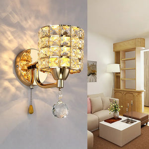 The Luxury Cylinder Wall Light with a crystal lampshade and pull chain is mounted on a white living room wall, complementing beige furniture and wooden shelves to bring luxurious, contemporary lighting to the space.