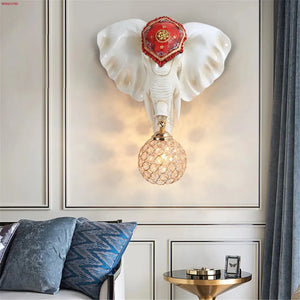 Transform your space with a unique ambiance using the 3D Elephant Wall Light. The elephant head, featuring a decorative red headpiece, supports a textured spherical light over the bed with blue pillows and a round side table.