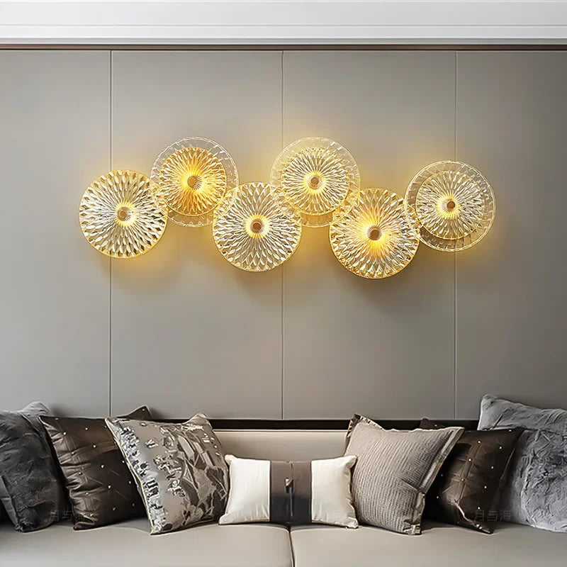 The Amber Deco Wall Sconce, featuring six circular textured glass panels, emits a warm glow above a grey sofa accented with assorted throw pillows.