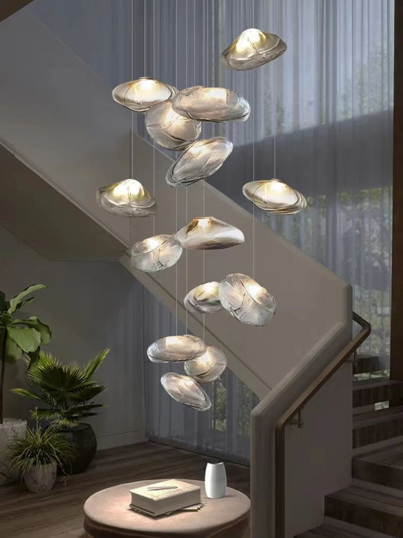 The Luxe Cloud Glass Pendant Light, with its sculptural cloud-like glass shades, hangs in the stairwell near a round table, indoor plant, and a large window draped with sheer curtains.