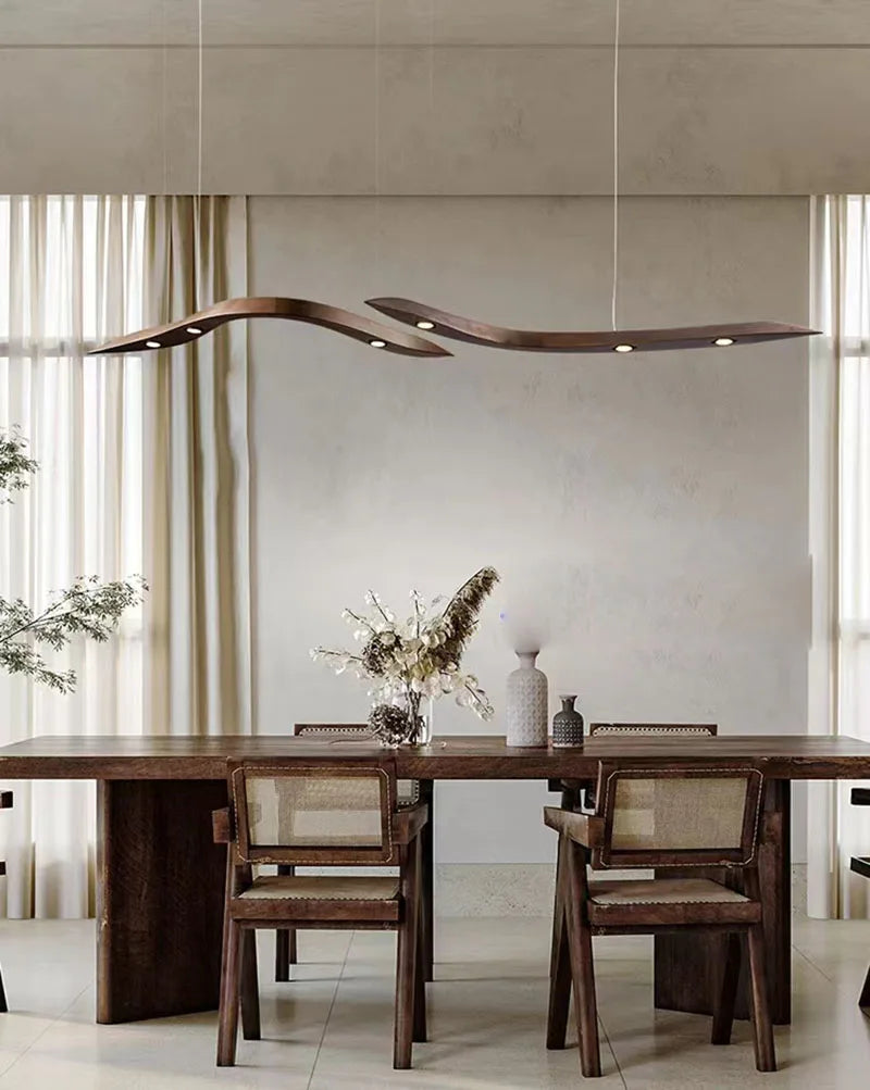 A modern dining room features a wooden table, four chairs, the Spiral Wood Pendant Light, and a vase of dried flowers; light curtains cover the windows in the background.