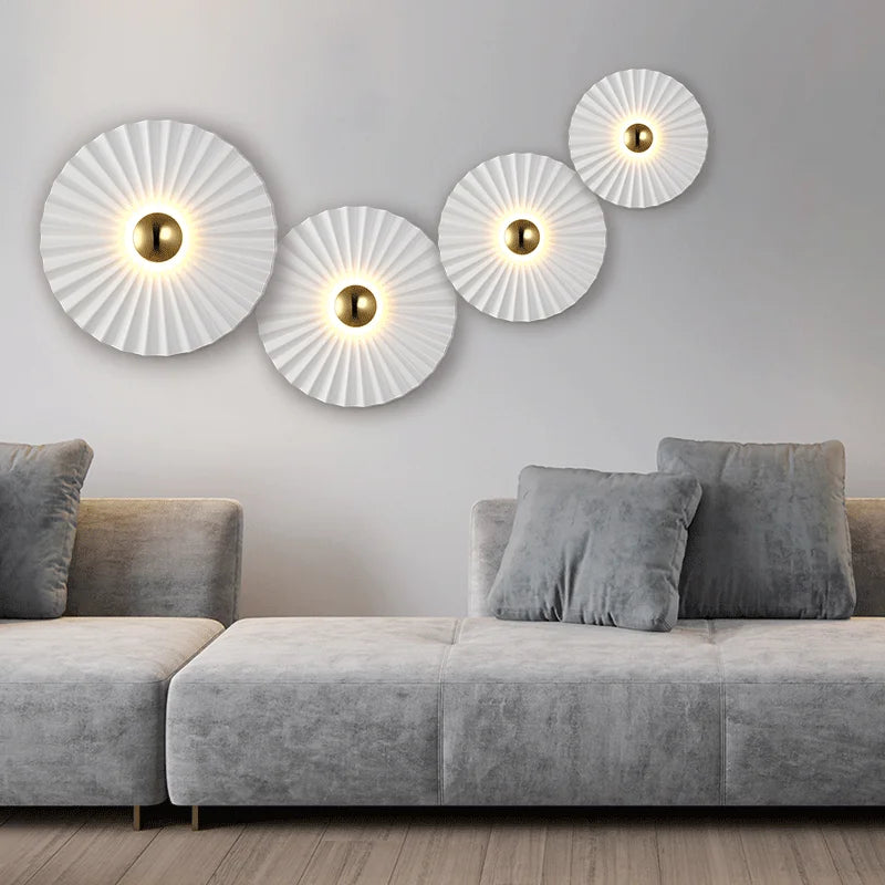 A modern living room features a grey sofa and four Origami Circle Wall Lights with pleated white surfaces and glowing centers, arranged diagonally on a light grey wall.