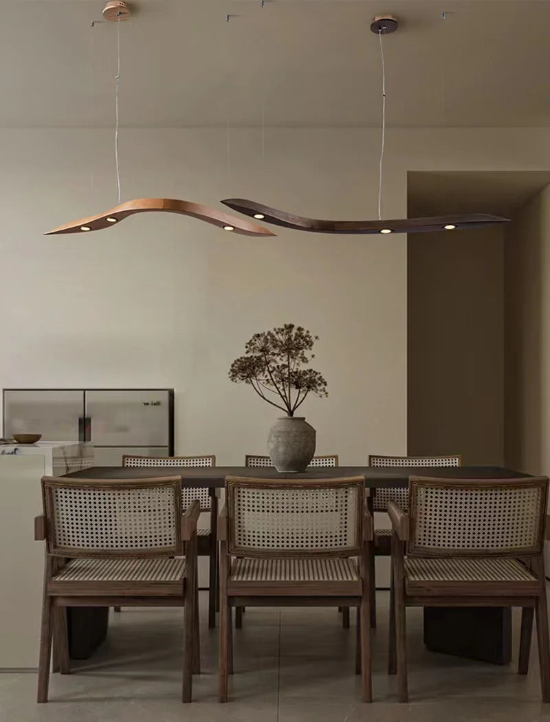 Dining area with a wooden table, six cane-backed chairs, a vase of dried flowers, and two Spiral Wood Pendant Lights—sleek, wavy fixtures—hanging overhead.