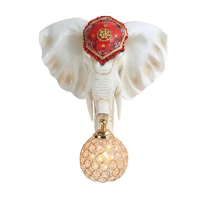 The 3D Elephant Wall Light has a red-embellished headpiece and holds a glowing round lampshade in its trunk, creating a unique ambiance.