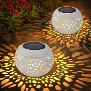 Two Solar Orb Lamps with intricate hollow iron cut-out patterns cast decorative yellow light along a garden path bordered by red flowers and lush green foliage at night.