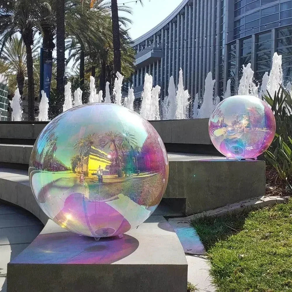Two large Inflatable Gazing Balls sit on concrete steps by a fountain, with palm trees and a modern glass building behind them, adding bold garden decor.