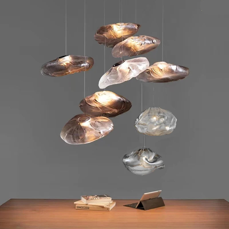 The Luxe Cloud Glass Pendant Light, with its sculptural cloud-inspired form, hangs above a wooden table holding an open book and tablet, standing out as a modern centerpiece against a plain gray background.