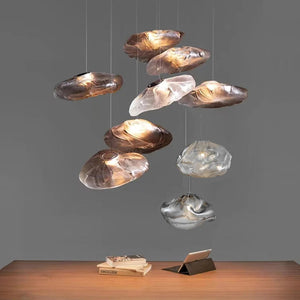 The Luxe Cloud Glass Pendant Light, with its sculptural cloud-inspired form, hangs above a wooden table holding an open book and tablet, standing out as a modern centerpiece against a plain gray background.