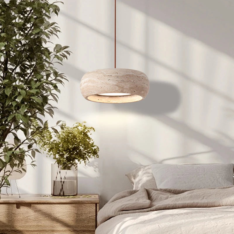 A modern bedroom features a dimmable Natural Stone Pendant Light, a wooden nightstand with green plants, and sunlight casting shadows on a white wall and an unmade bed.
