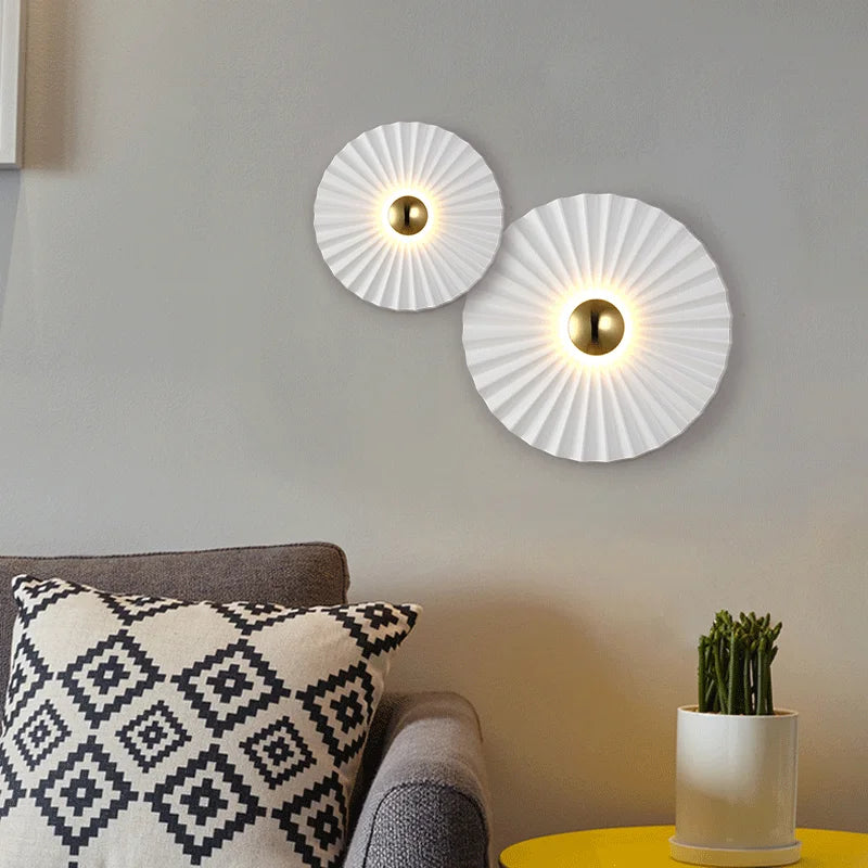 Two Origami Circle Wall Lights are mounted on a grey wall above a sofa with a geometric-patterned pillow and a yellow table holding a small potted plant.