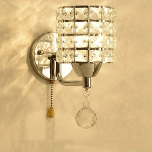 The Luxury Cylinder Wall Light features a crystal-patterned shade, metal base, pull chain, and hanging crystal ornament to add luxury lighting and elegance to your space, beautifully showcased on a beige wall.