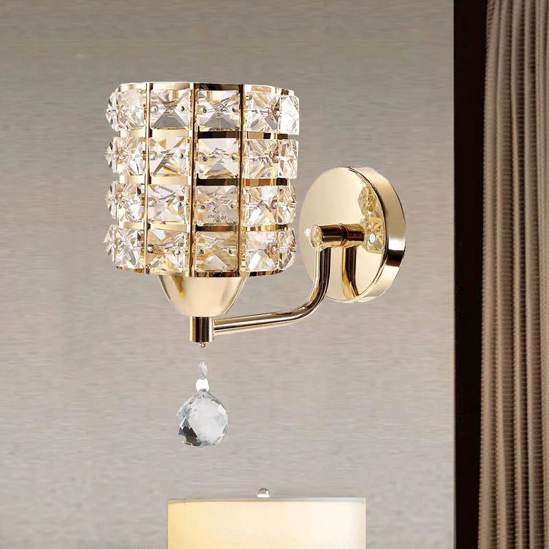 The Luxury Cylinder Wall Light is a gold crystal wall light with a grid-patterned shade and single hanging crystal, mounted on a beige wall next to a curtain.