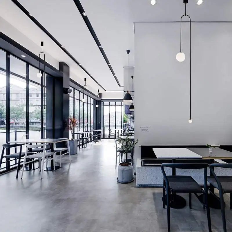 A modern café interior with large windows and minimalist furniture features sculptural lighting, including Minimalist Line Lights. Sparse greenery adds a natural touch, enhanced by modern lighting that complements the ambiance perfectly.
