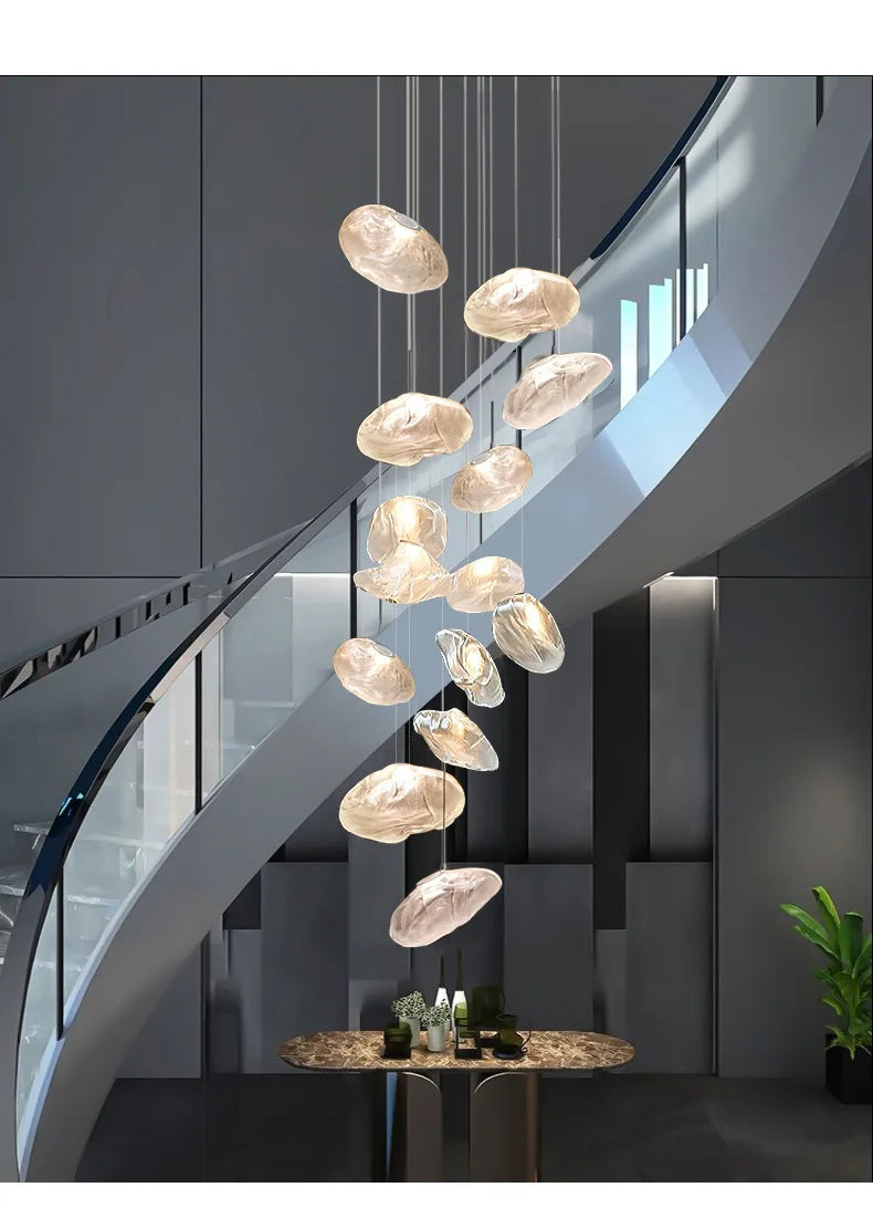 The Luxe Cloud Glass Pendant Lights hang in front of a curved staircase with glass railings, illuminating a small table adorned with drinks and greenery in a contemporary interior.