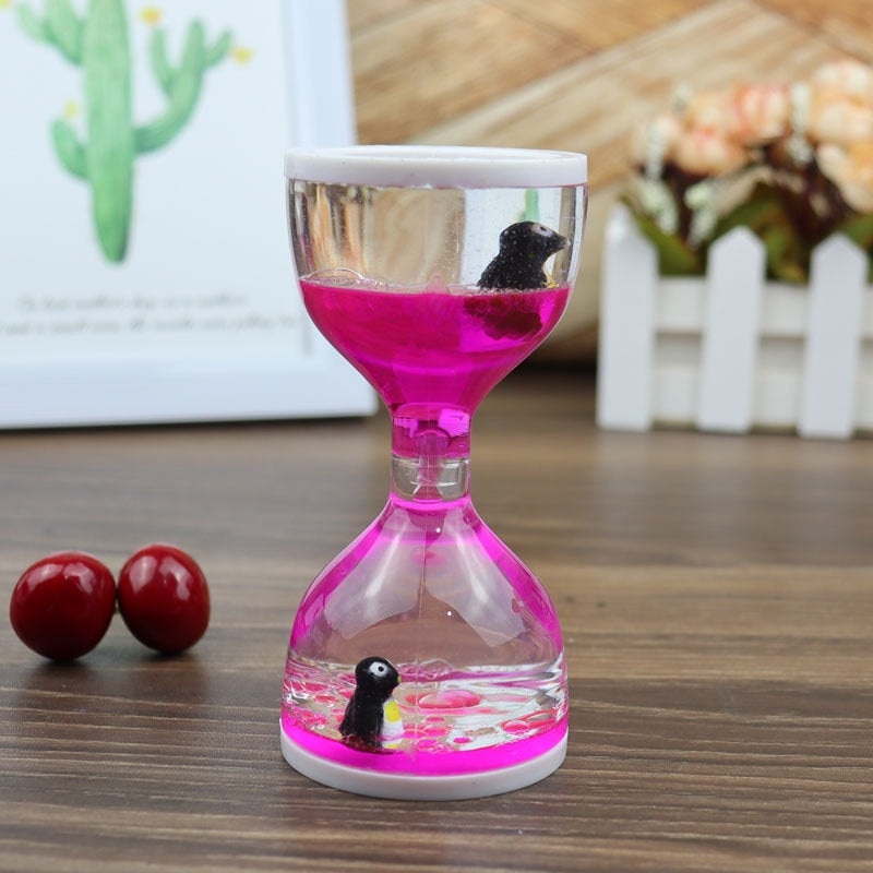 The visually stunning Dual Color Liquid Hourglass features a striking display of pink liquid and tiny black figures encased within it, all set on a wooden surface. Beside the hourglass are small décor items and scattered red beads, while a framed cactus image is softly blurred in the background.