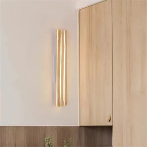 The Rectangle Wood Wall Light, a minimalist LED fixture with a gold vertical sconce, emits a warm ambient glow and is smoothly mounted on a neutral wall, enhancing the light wooden cabinets’ aesthetic.