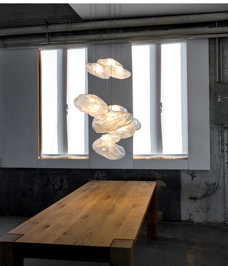 A wooden table sits beneath the Luxe Cloud Glass Pendant Light, shaped like clouds, in front of two tall windows draped with sheer white curtains in an industrial-style room.