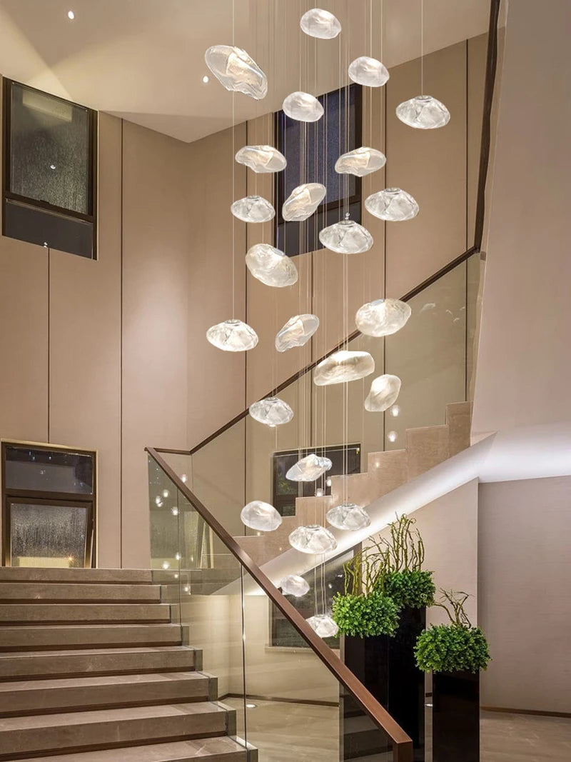 The Luxe Cloud Glass Pendant Light, featuring textured glass and multiple pendants, hangs in a spacious foyer with a staircase, glass railing, and potted plants.