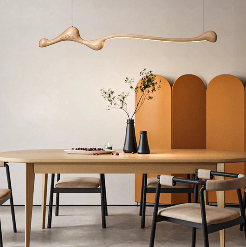 Minimalist dining room with a wooden table, four chairs, the Tideform Biomorphic Pendant Light overhead, two black vases with branches, and an orange folding screen set against a neutral wall.