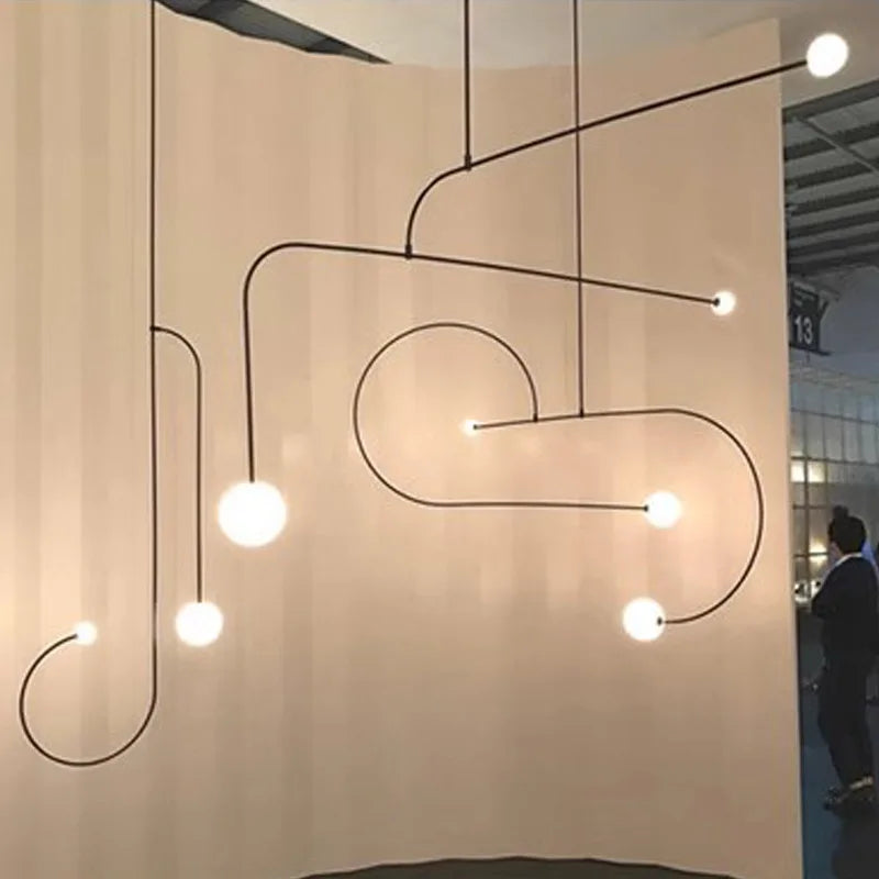 A modern art installation features the Minimalist Line Light with twisted black rods and spherical bulbs set against a beige backdrop. Nearby, a person observes this captivating indoor display.