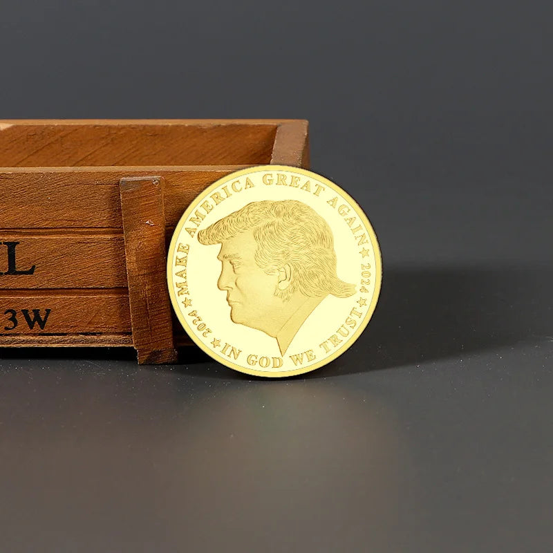 The Trump Commemorative Coin, gold-colored and featuring a profile image with "MAKE AMERICA GREAT AGAIN 2024" and "IN GOD WE TRUST," leans against a wooden box from the Save America Legacy Collection.