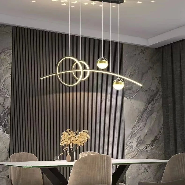 A modern dining room features a marble wall, ribbed paneling, and geometric illumination from the sleek Stellar Pendant Light, which is hung above a table with beige chairs and a vase of dried plants.
