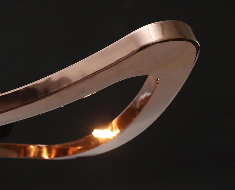 A close-up of the Elegant Industrial Gallery Pendant, featuring a metallic rose gold finish, an abstract sculptural shape, and a smooth, reflective surface, set against a black background.