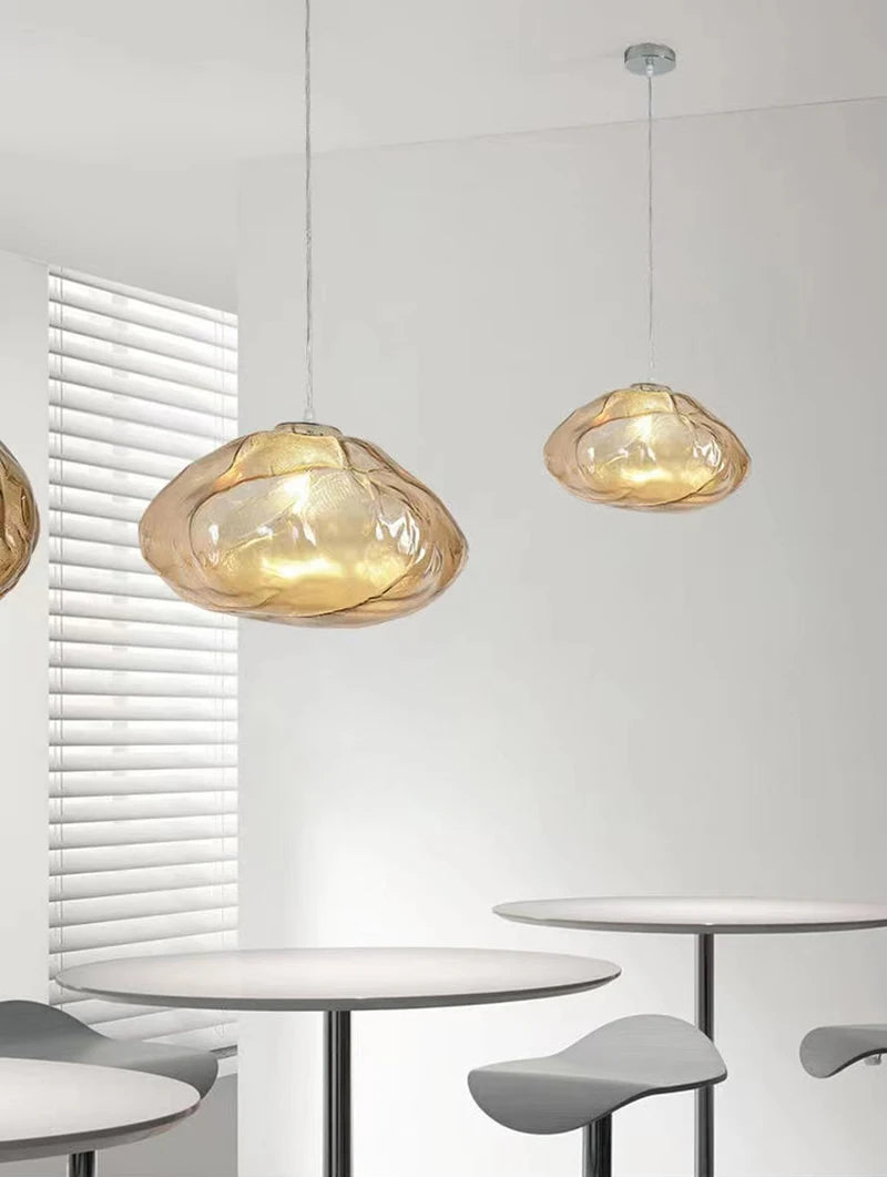 Two Luxe Cloud Glass Pendant Lights with translucent amber shades hang above round white tables and gray stools in a minimalist room with white walls and window blinds.