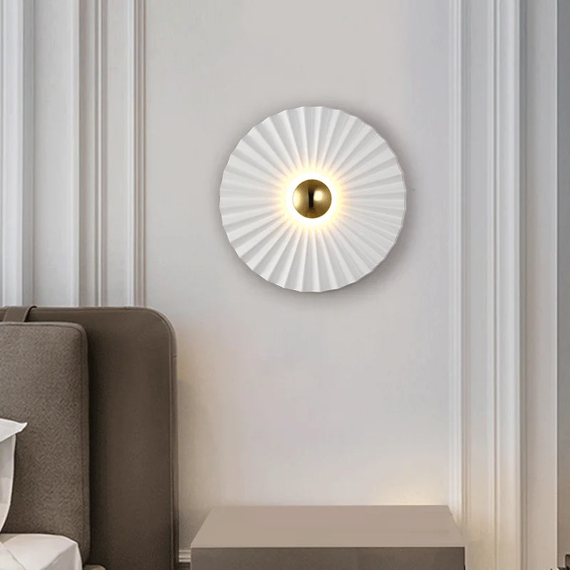 The Origami Circle Wall Light, featuring a modern pleated white shade and central gold accent, is mounted above a bedside table beside a bed.