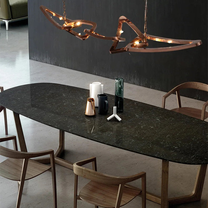 A modern dining table with a dark, oval stone top and wooden chairs is illuminated by the Elegant Industrial Gallery Pendant above, with decorative objects placed on the tabletop.