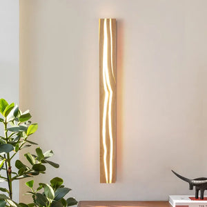 The Rectangle Wood Wall Light, designed with a minimalist and vertical look, illuminates a plain wall with its wavy design. A green plant in the corner enhances the scene, while a wooden table with books complements the organic aesthetic.