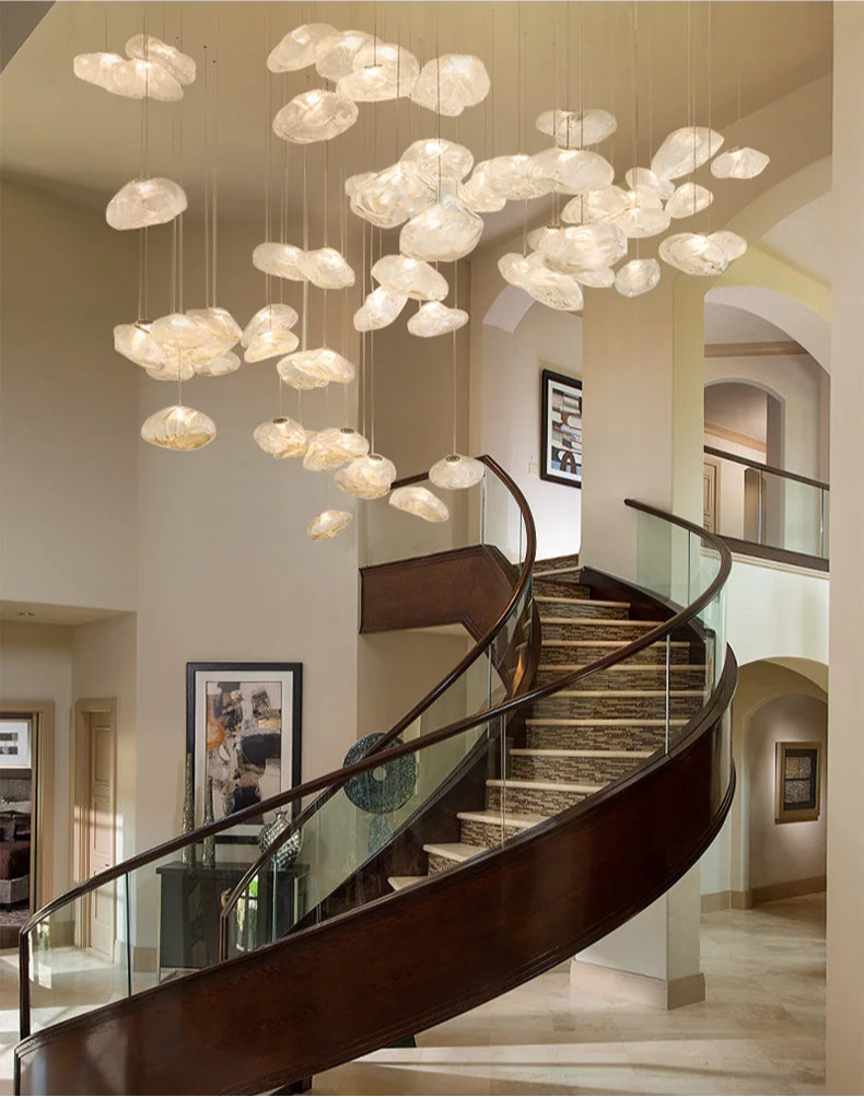 A modern spiral staircase with glass railing stands beneath a ceiling installation featuring Luxe Cloud Glass Pendant Lights—sculptural, translucent disc-shaped lamps—in a spacious, elegant interior.