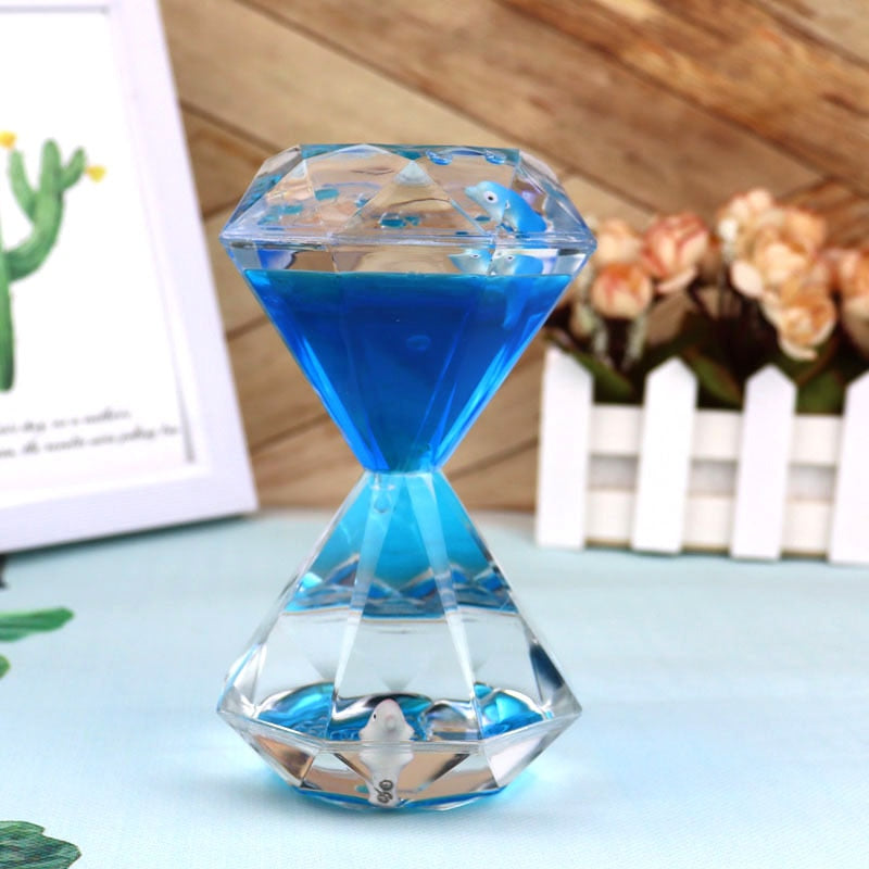 A Dual Color Liquid Hourglass, with its visually stunning diamond-shaped design, rests on a light blue tablecloth. It is surrounded by a wooden photo frame, a green plant, and a white flower pot filled with pink flowers.