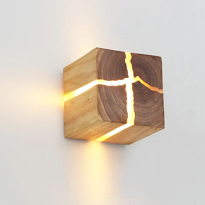 The Wood Cube Wall Light highlights its natural wood grain, casting a warm glow through delicate openings when mounted on a light-colored wall.