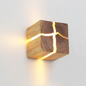 The Wood Cube Wall Light highlights its natural wood grain, casting a warm glow through delicate openings when mounted on a light-colored wall.