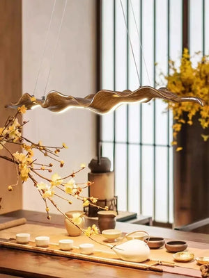 The Leaf Pendant Light, with its organic-inspired, sculptural design, hangs over a wooden dining table set featuring teapots, cups, and decorative branches adorned with yellow flowers—ideal for modern spaces.