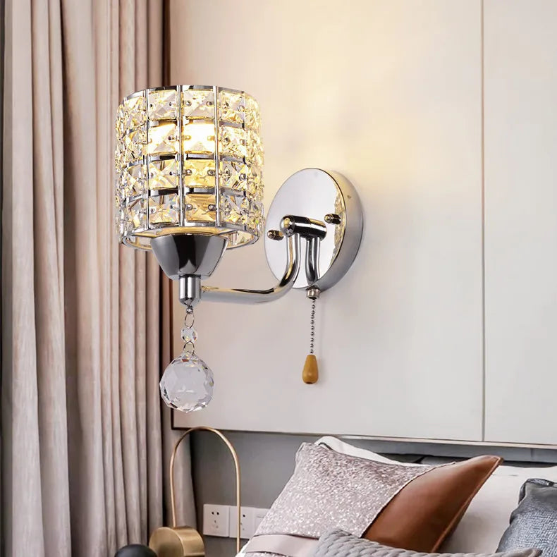 The Luxury Cylinder Wall Light with a pull chain switch is mounted above a bed with cushions and a side table, set against light walls and curtains, bringing elegant illumination to the serene room.