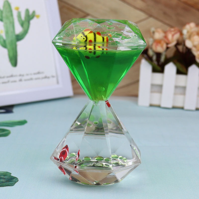 The Dual Color Liquid Hourglass features a green liquid-filled, hourglass-shaped container with a dual-color liquid display showcasing a decorative yellow and red striped object inside. The background includes a framed image of a cactus and some flowers in a white picket fence pot, creating a visually stunning experience.